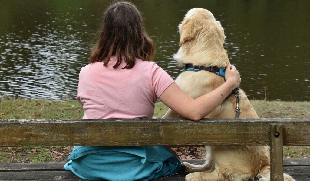 A woman in a pink shirt sits on a wooden bench by a lake with her golden retriever, her arm wrapped around the dog in a moment of companionship.