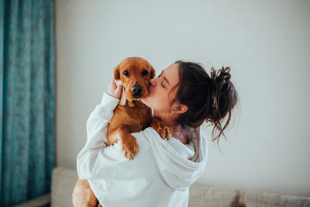 A woman in a white hoodie kissing a brown dog she is holding, with a cozy indoor background.