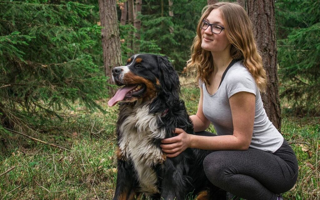 A woman with glasses and wavy brown hair kneels beside a large black, white, and tan dog in a forest. The dog is panting happily, and the two share a peaceful moment surrounded by tall trees and lush greenery.