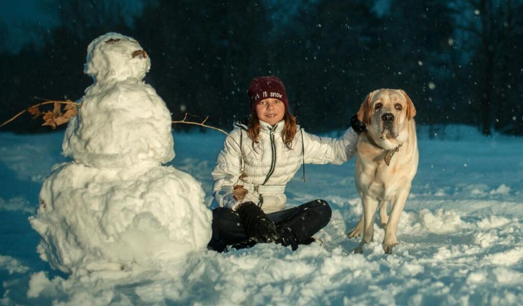 Child sitting with a dog next to a snowman.