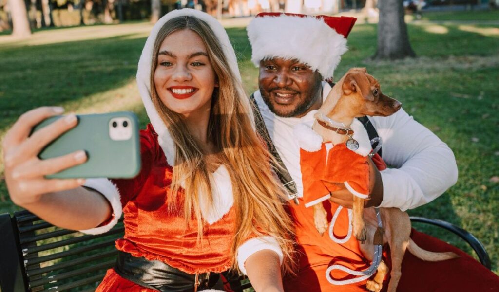 Two people in Santa outfits taking a selfie with a small dog.