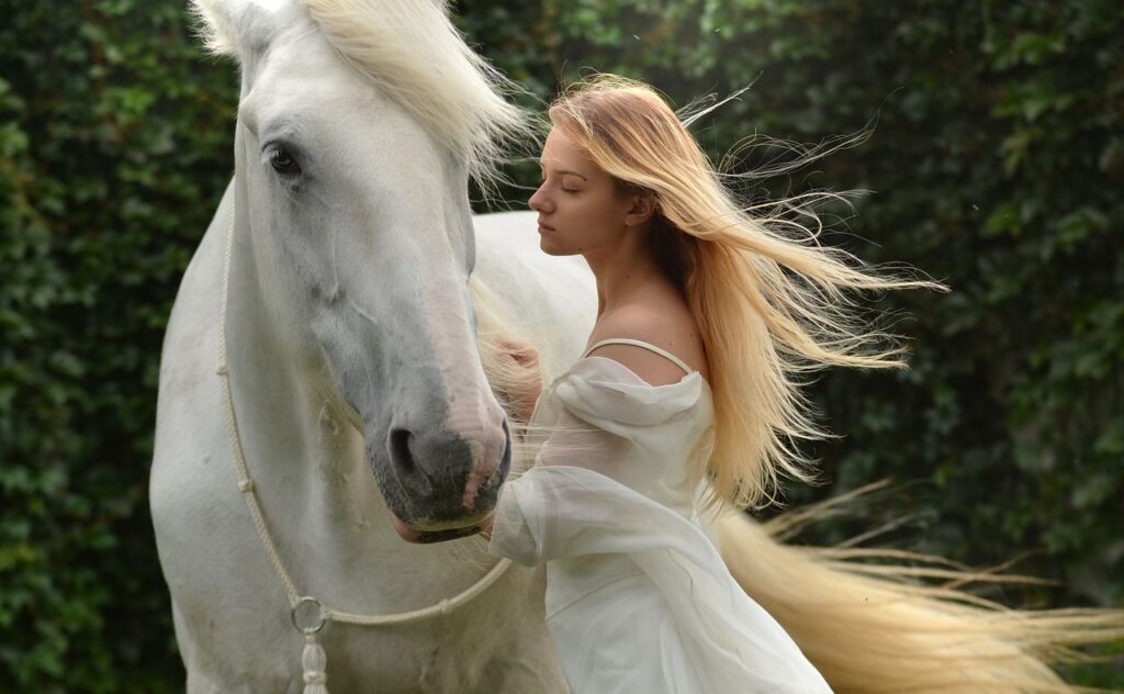 A young woman with flowing blonde hair stands close to a white horse, both exuding calmness in a lush green outdoor setting.
