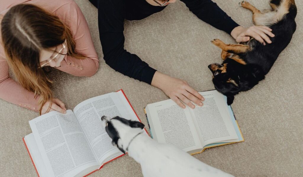 Two people reading books on the floor, with two dogs nearby—one relaxing and the other curious.