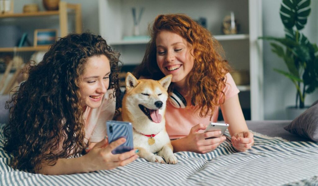 Two smiling women with a Shiba Inu dog on a bed.