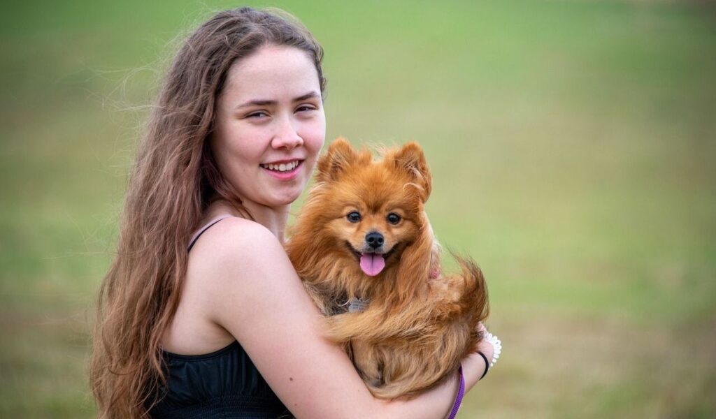 A smiling woman with long brown hair holds a small, fluffy orange dog. The dog has its tongue out and a cheerful expression, standing out against a blurred green background.