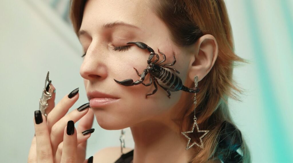 Close-up of a woman with a black scorpion on her cheek, wearing star earrings and black nails.