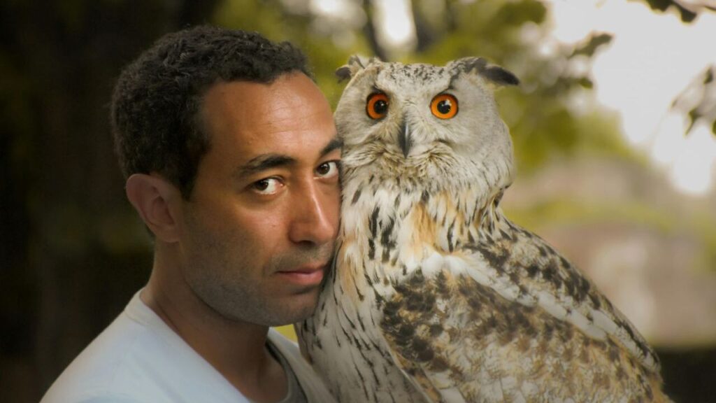 A man closely interacting with a majestic owl, emphasizing a unique bond between human and wildlife, with the owl's vibrant orange eyes standing out vividly against its patterned feathers
