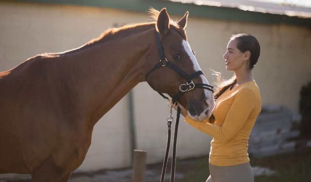 A woman in a yellow top gently holding the reins of a brown horse while looking at it.
