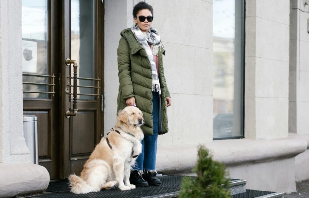 Woman in a green coat stands with her service dog outside a building.