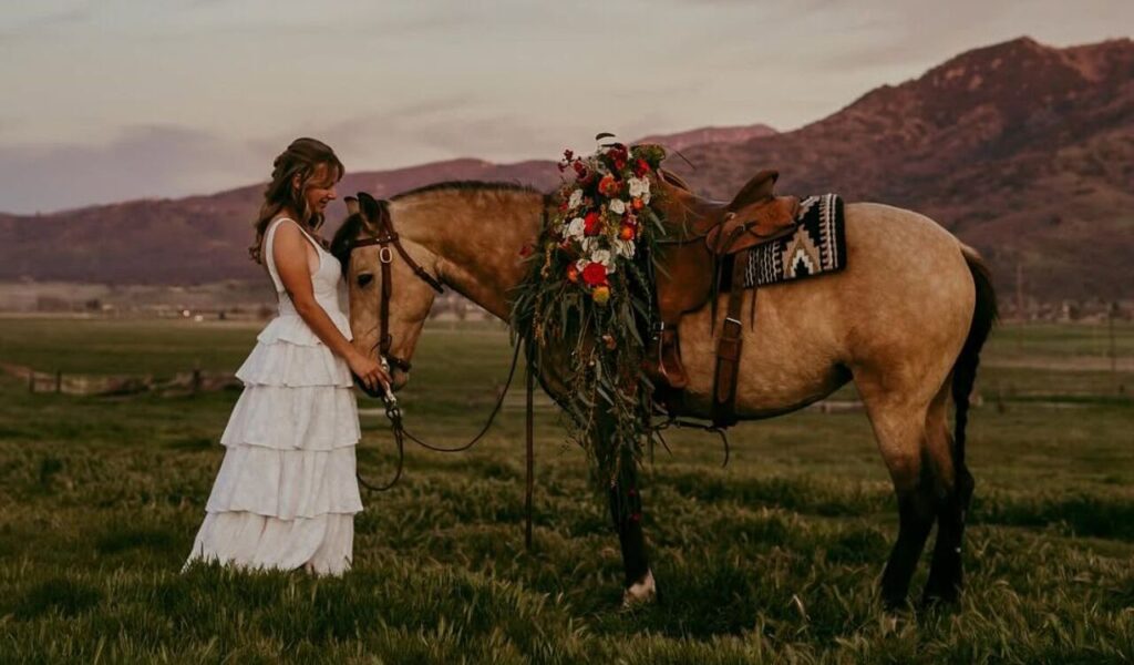 A woman in a white dress stands gently with a decorated wild horse in a peaceful meadow